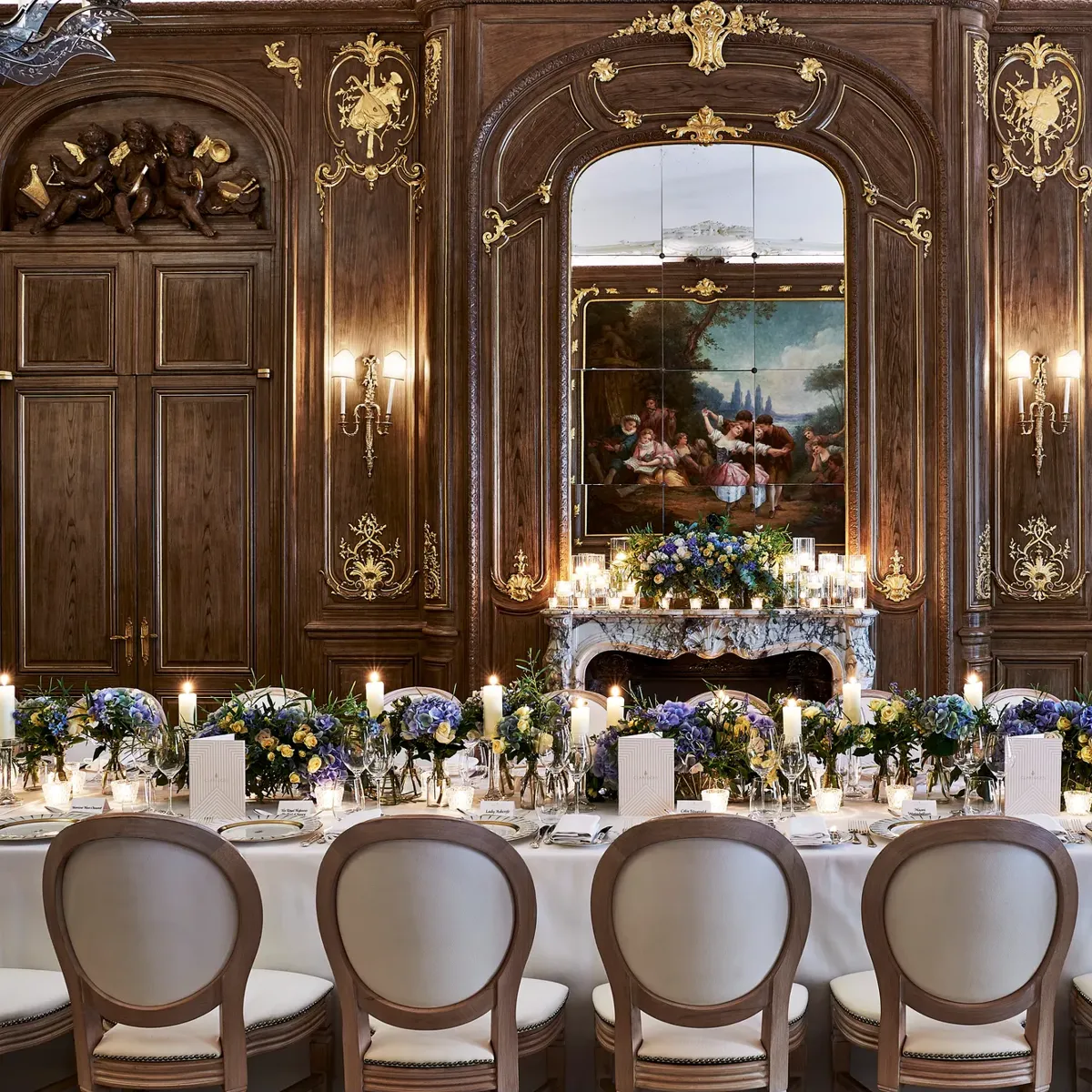 Grand candlelit banquet table with blue and white florals set before ornate wood-panelled walls, gilded details, and a classical painting.