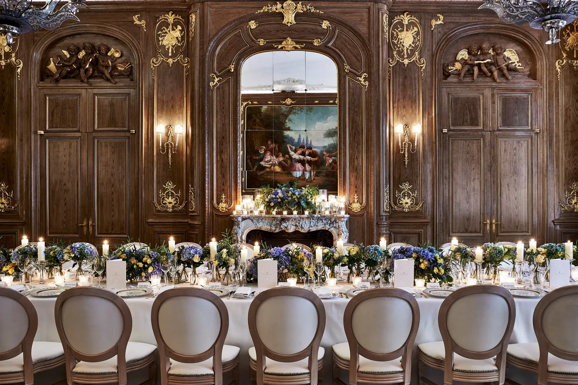 Grand candlelit banquet table with blue and white florals set before ornate wood-panelled walls, gilded details, and a classical painting.