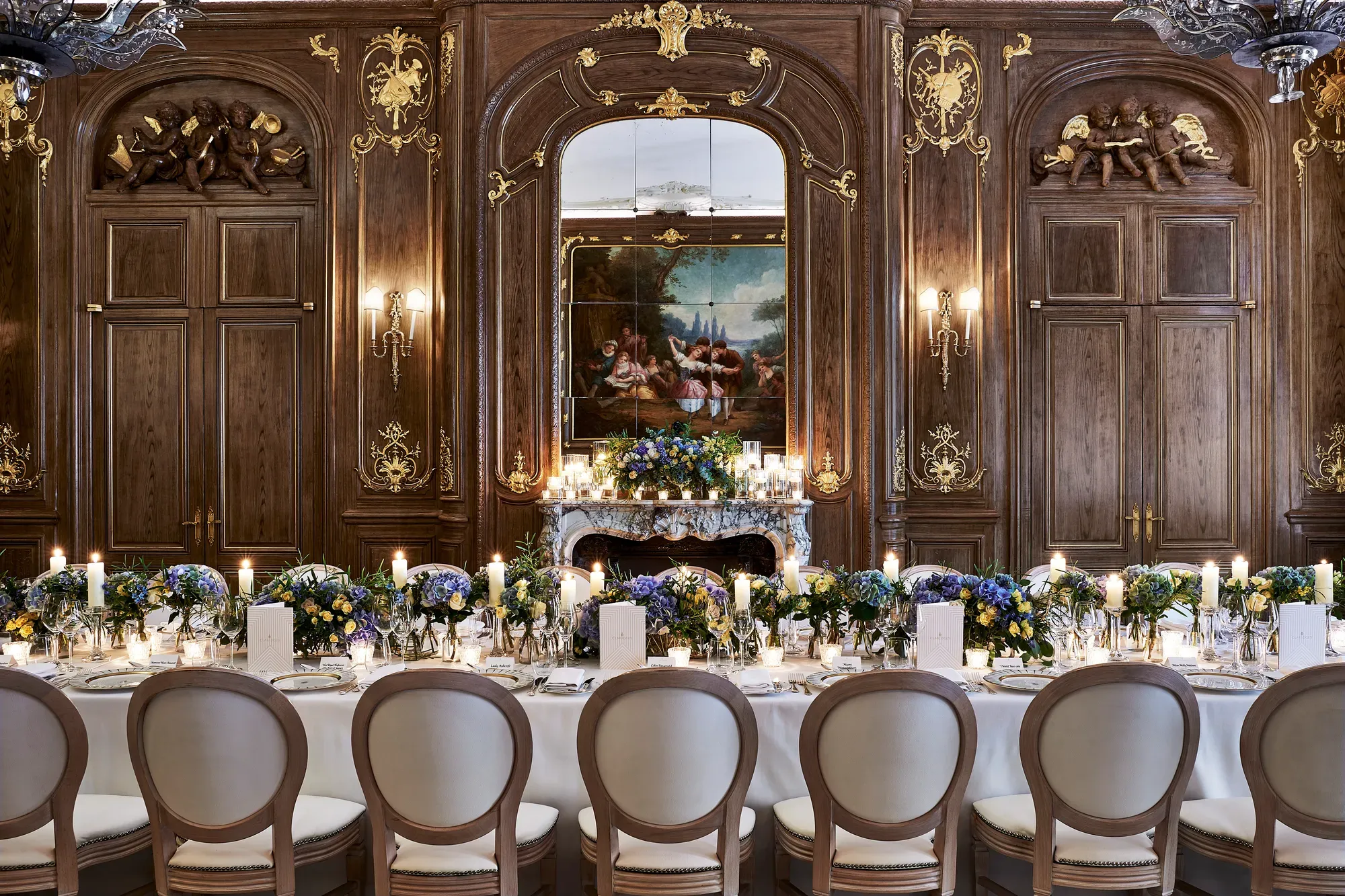 Grand candlelit banquet table with blue and white florals set before ornate wood-panelled walls, gilded details, and a classical painting.