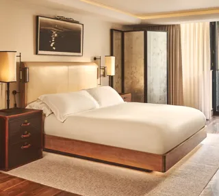 Warm, modern bedroom with a cream upholstered bed, wooden furniture, soft lighting, and sheer curtains leading to a balcony door.