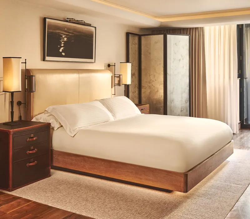 Warm, modern bedroom with a cream upholstered bed, wooden furniture, soft lighting, and sheer curtains leading to a balcony door.