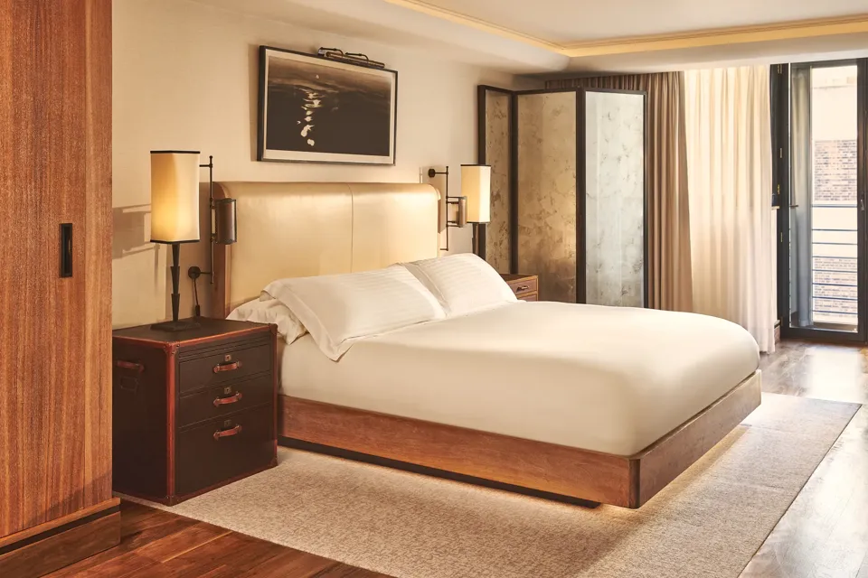 Warm, modern bedroom with a cream upholstered bed, wooden furniture, soft lighting, and sheer curtains leading to a balcony door.