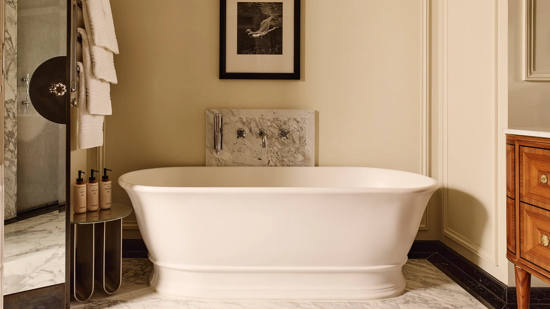 Elegant bathroom featuring a freestanding white bathtub with marble accents, a framed black-and-white photograph above, and soft lighting that highlights the cream walls and wood cabinetry.