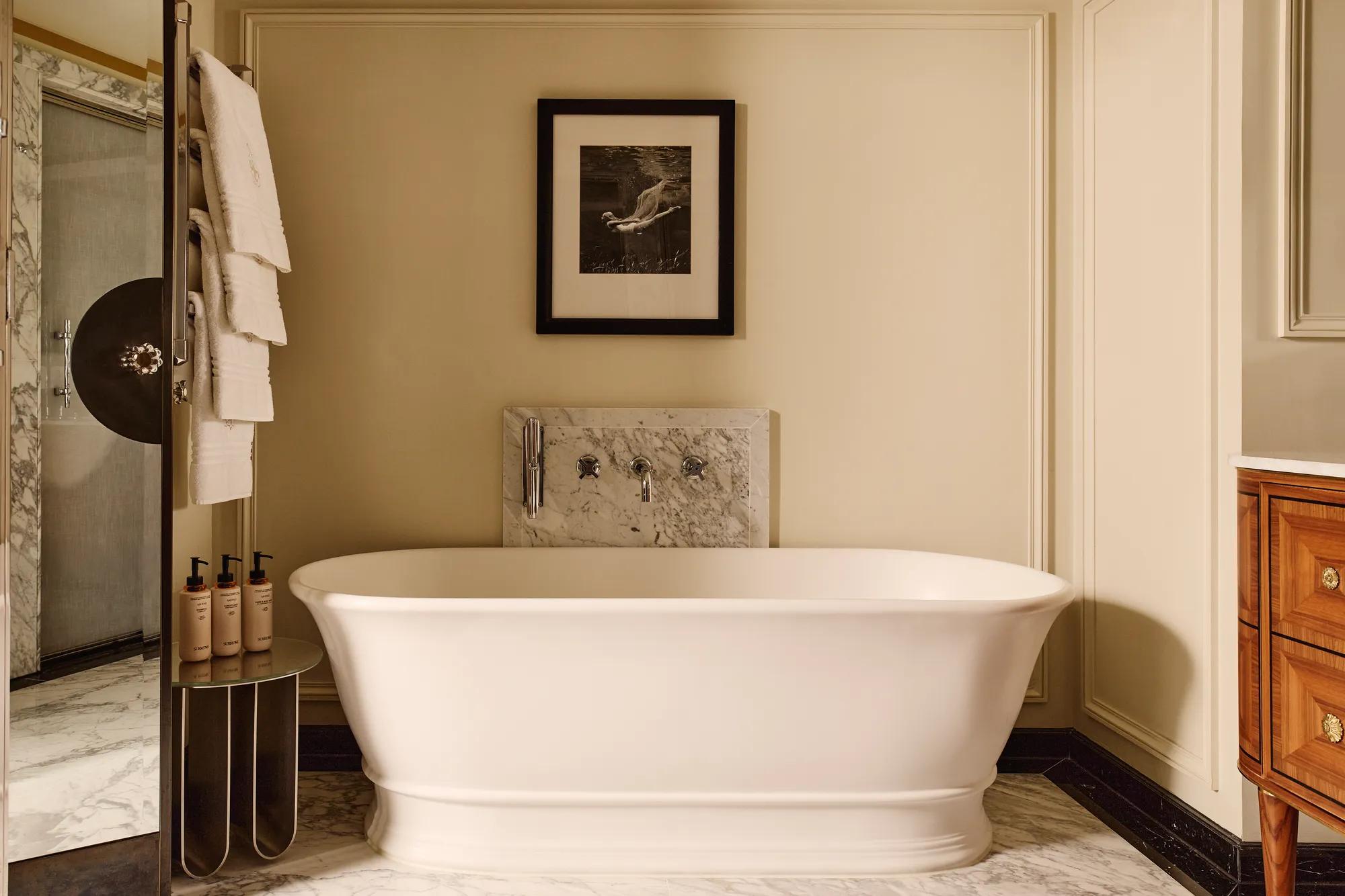 Elegant bathroom featuring a freestanding white bathtub with marble accents, a framed black-and-white photograph above, and soft lighting that highlights the cream walls and wood cabinetry.
