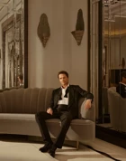 Man in tuxedo sits on curved sofa in elegant mirrored lounge, with art deco wall lights and warm, ambient lighting.
