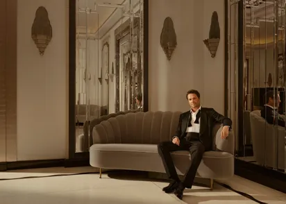 Man in tuxedo sits on curved sofa in elegant mirrored lounge, with art deco wall lights and warm, ambient lighting.