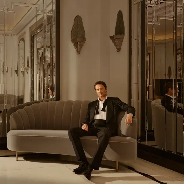 Man in tuxedo sits on curved sofa in elegant mirrored lounge, with art deco wall lights and warm, ambient lighting.