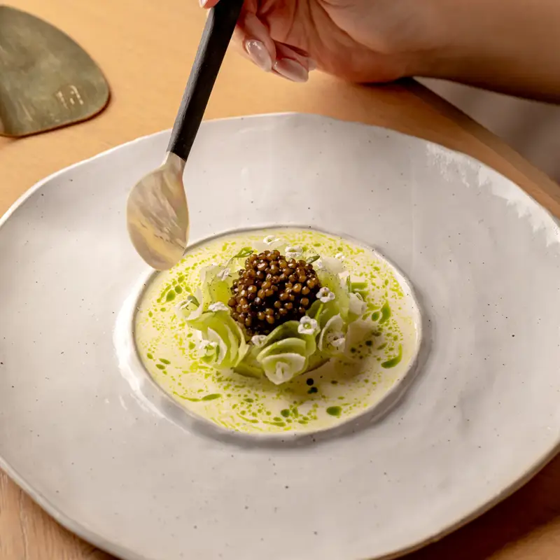 Hand holding a spoon over a plated dish with caviar, sliced vegetables and green sauce in a shallow bowl