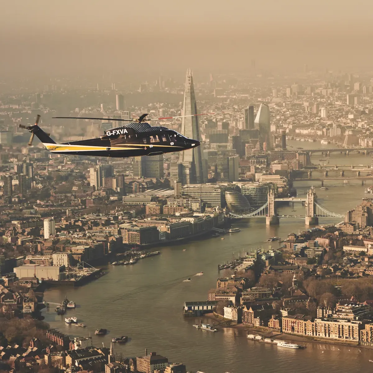 Helicopter flying over the River Thames with Tower Bridge, The Shard, and London skyline visible in the background.
