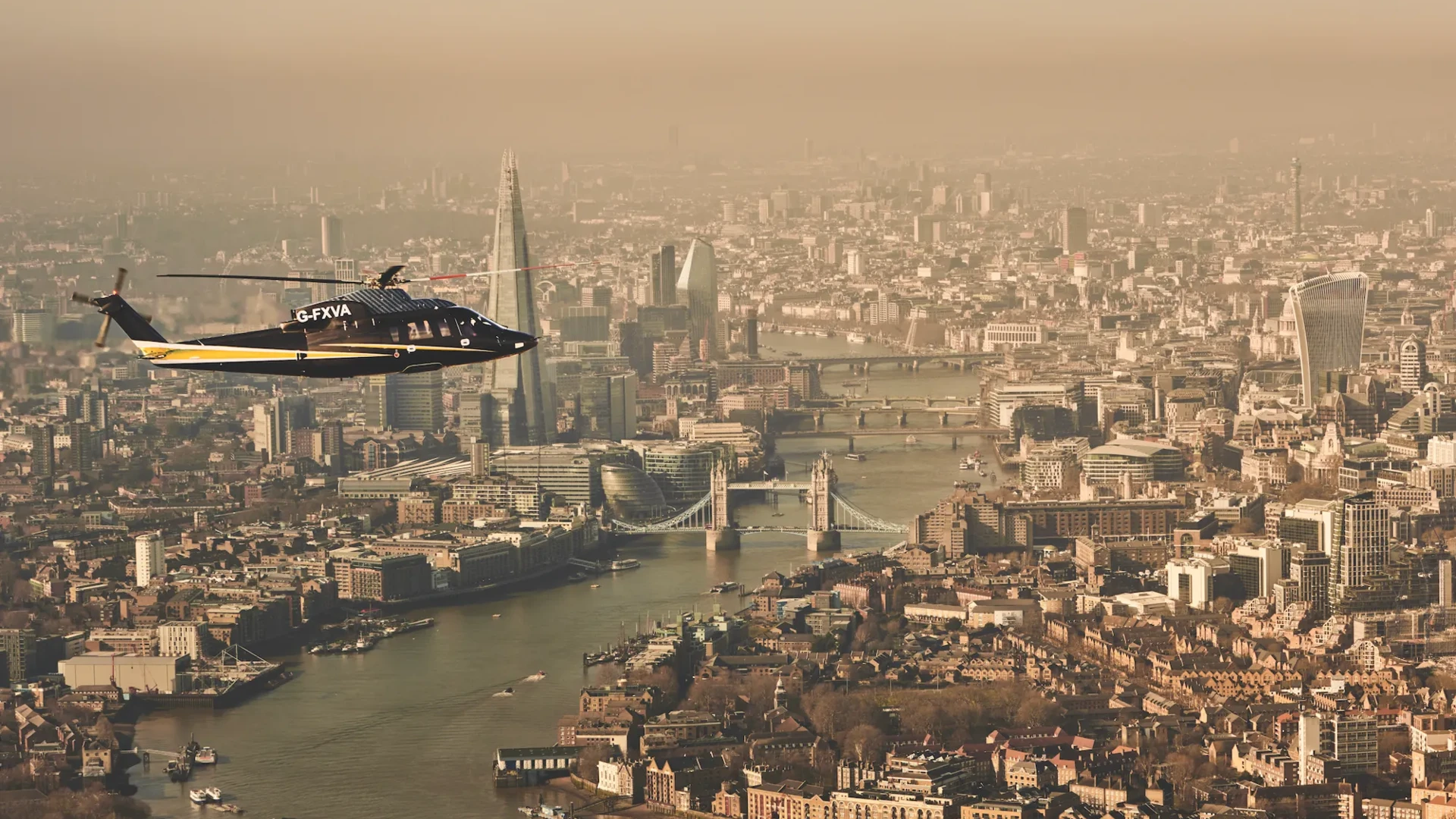 Helicopter flying over the River Thames with Tower Bridge, The Shard, and London skyline visible in the background.