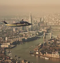 Helicopter flying over the River Thames with Tower Bridge, The Shard, and London skyline visible in the background.
