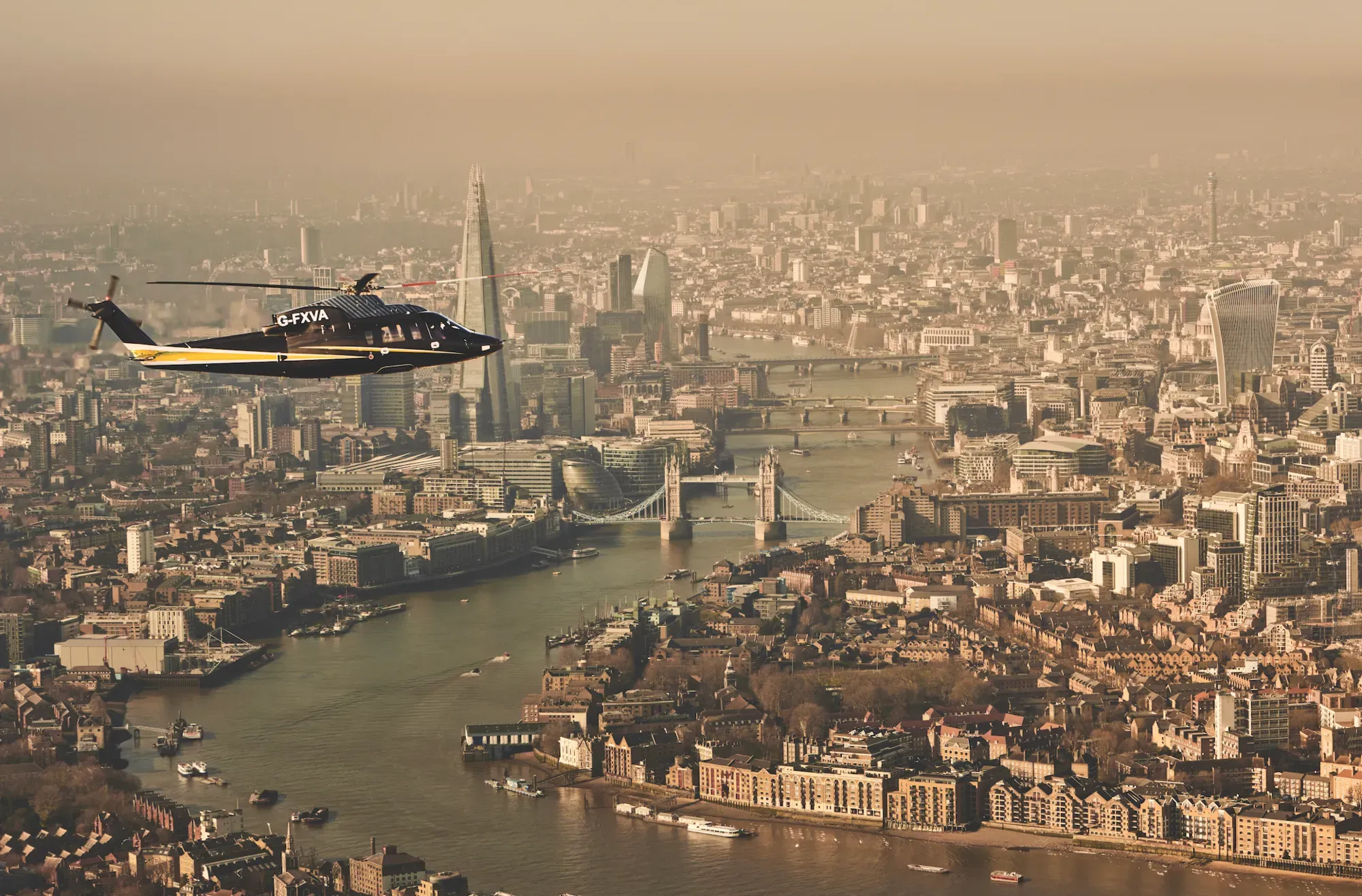 Helicopter flying over the River Thames with Tower Bridge, The Shard, and London skyline visible in the background.