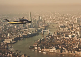 Helicopter flying over the River Thames with Tower Bridge, The Shard, and London skyline visible in the background.