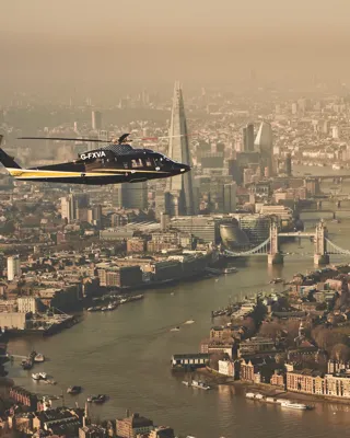 Helicopter flying over the River Thames with Tower Bridge, The Shard, and London skyline visible in the background.