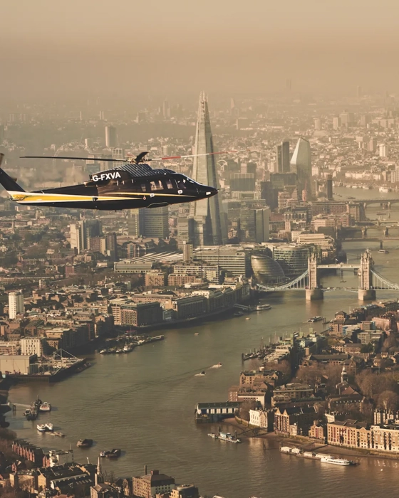 Helicopter flying over the River Thames with Tower Bridge, The Shard, and London skyline visible in the background.