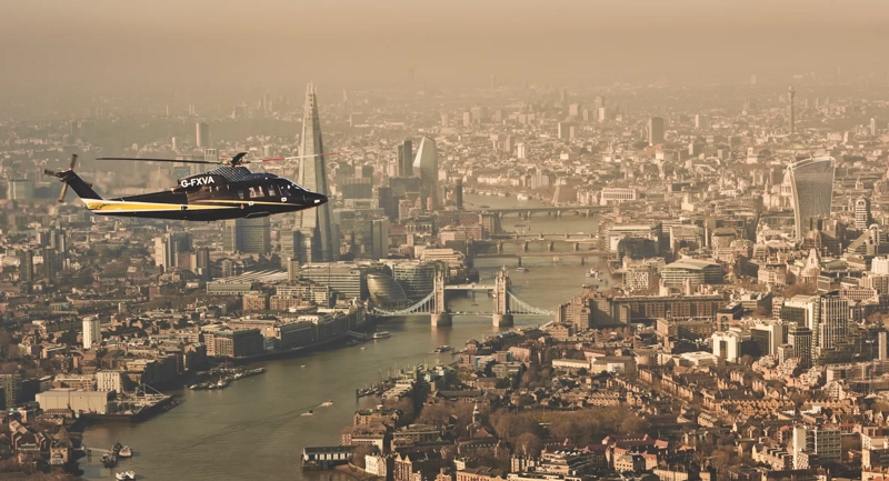 Helicopter flying over the River Thames with Tower Bridge, The Shard, and London skyline visible in the background.