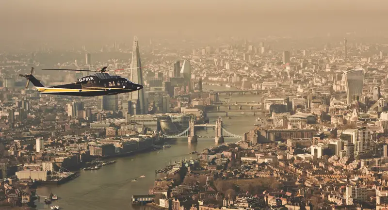 Helicopter flying over the River Thames with Tower Bridge, The Shard, and London skyline visible in the background.