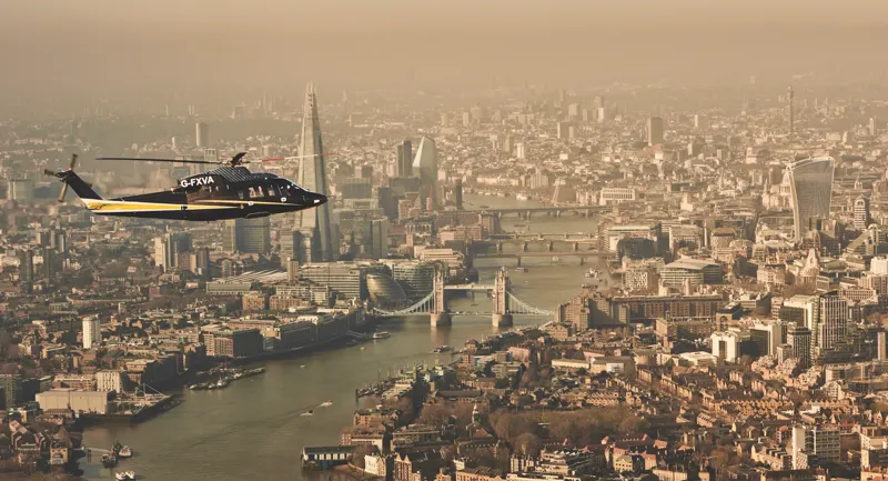 Helicopter flying over the River Thames with Tower Bridge, The Shard, and London skyline visible in the background.