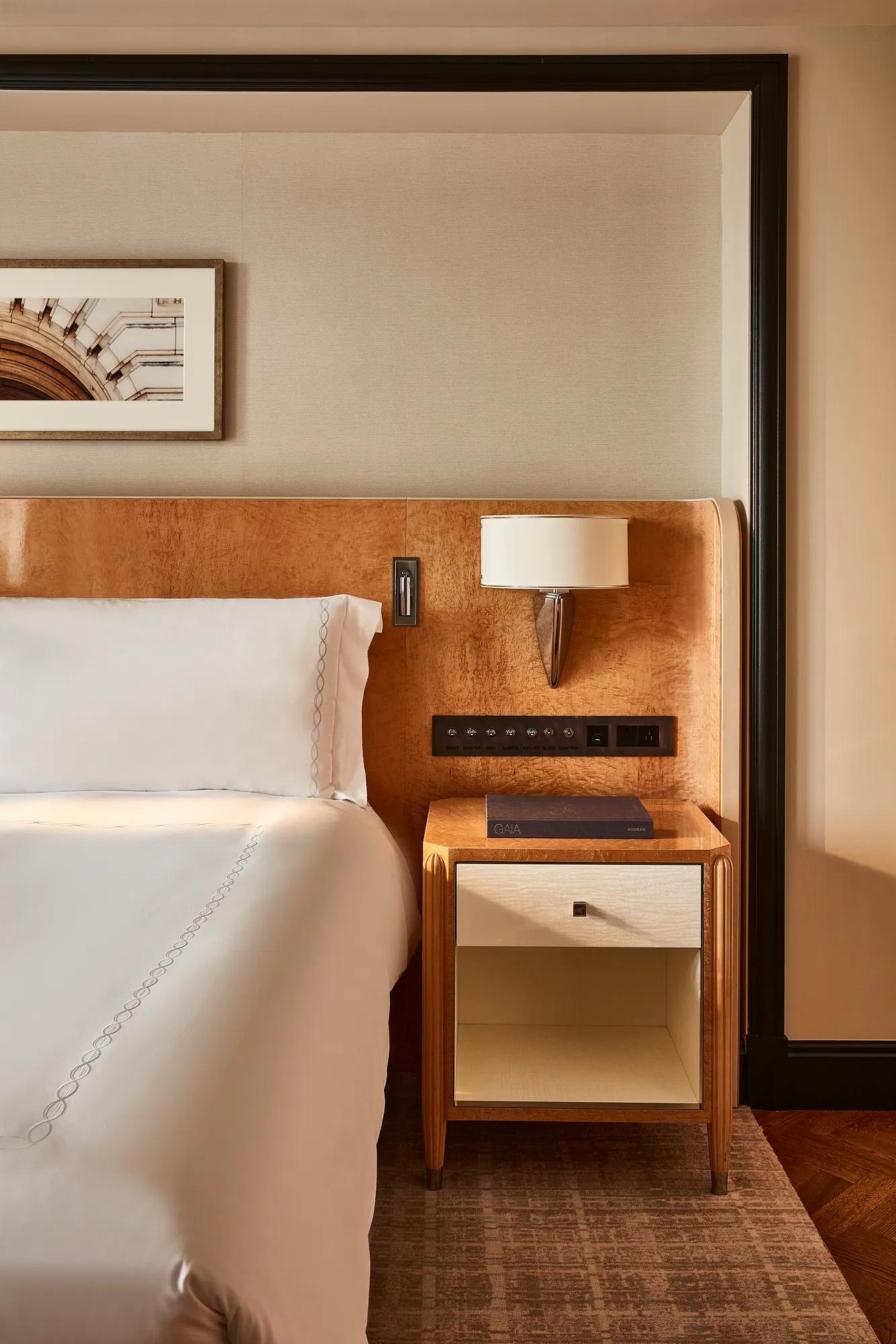 Bed with white linens and wood headboard, bedside table with drawer and book, wall-mounted lamp, and framed artwork on neutral wall