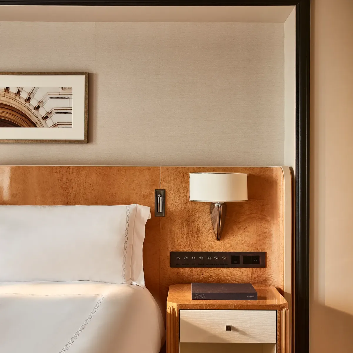 Bed with white linens and wood headboard, bedside table with drawer and book, wall-mounted lamp, and framed artwork on neutral wall
