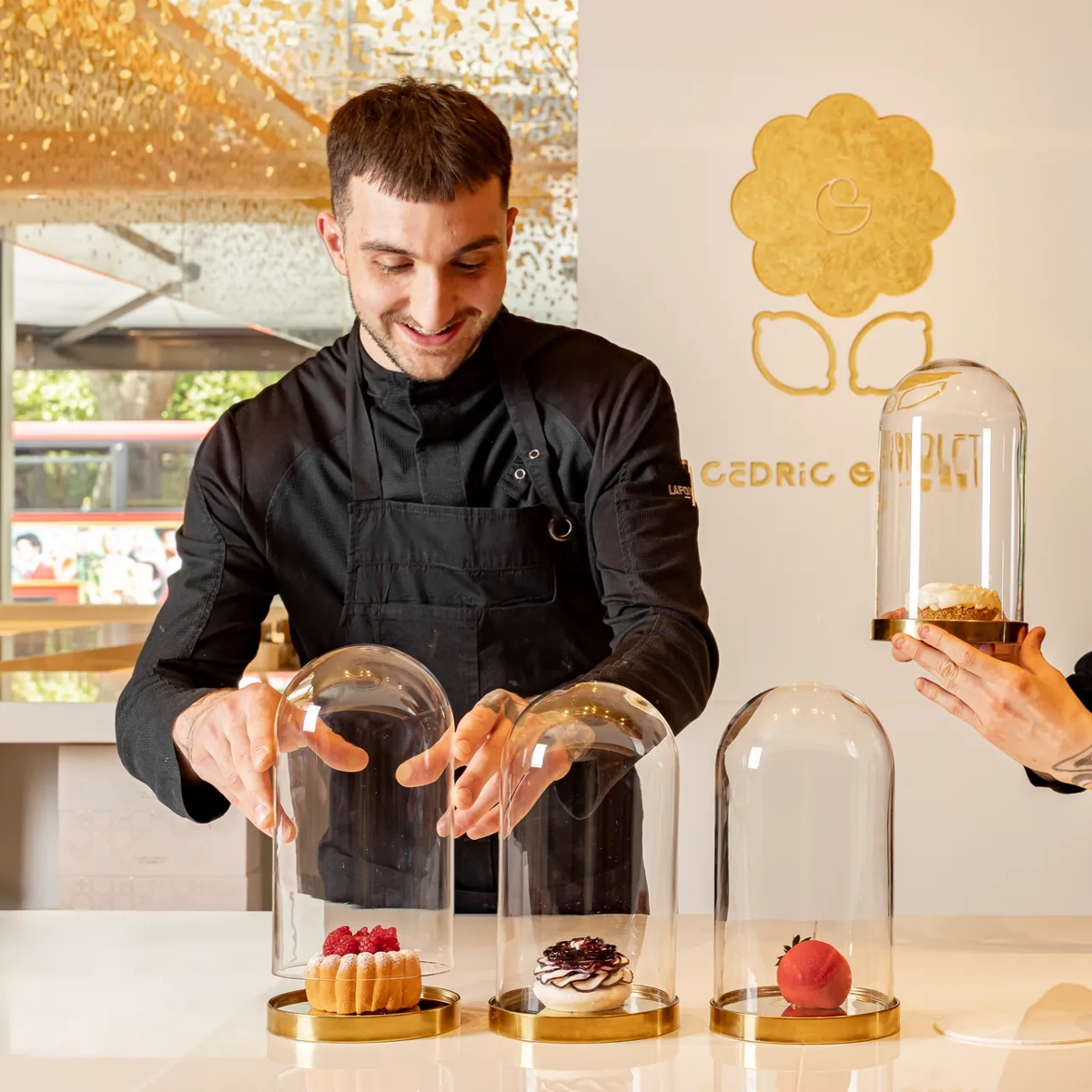 Pastry chef presenting desserts beneath glass domes on a counter in a bright pâtisserie setting.