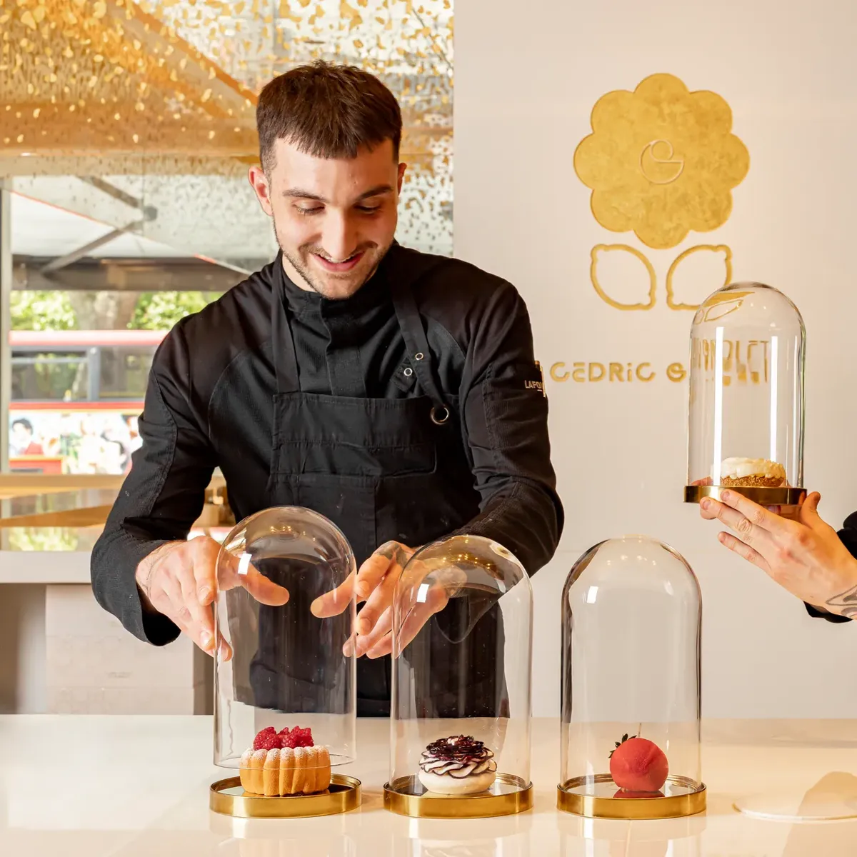 Pastry chef presenting desserts beneath glass domes on a counter in a bright pâtisserie setting.