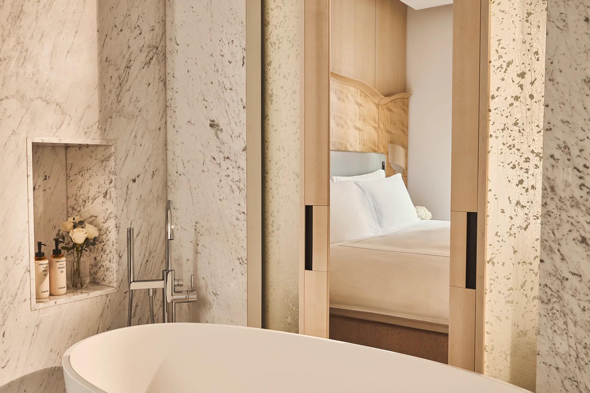 Freestanding bathtub beside marble wall with built-in shelf and view through sliding doors to bedroom with wood headboard.