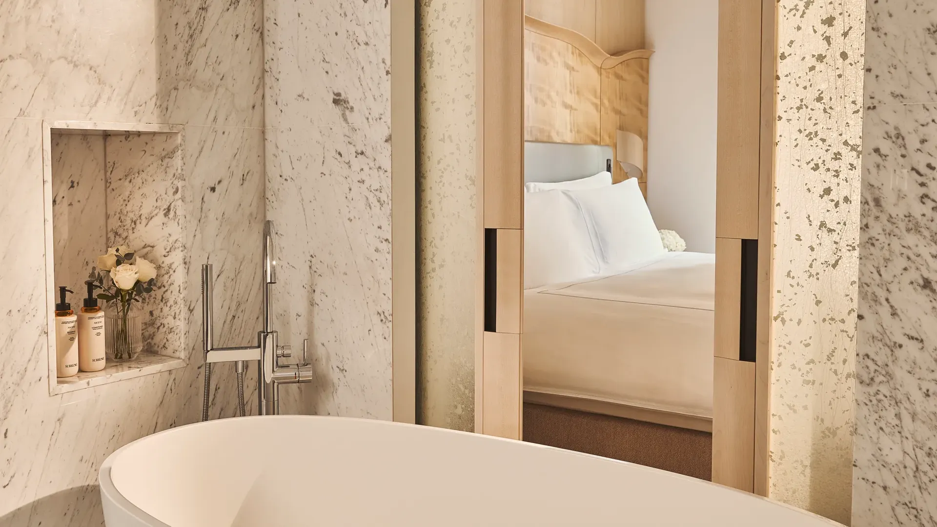 Freestanding bathtub beside marble wall with built-in shelf and view through sliding doors to bedroom with wood headboard.