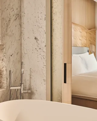 Freestanding bathtub beside marble wall with built-in shelf and view through sliding doors to bedroom with wood headboard.