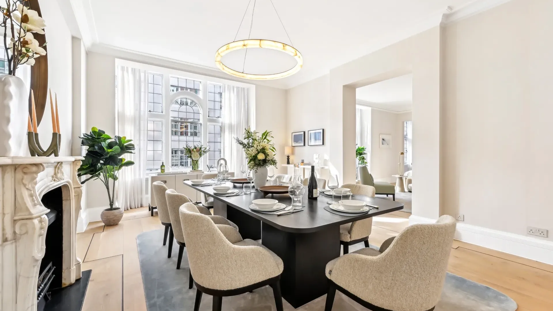 Airy dining room with a long dark table, cream chairs, chandelier, and large arched window.