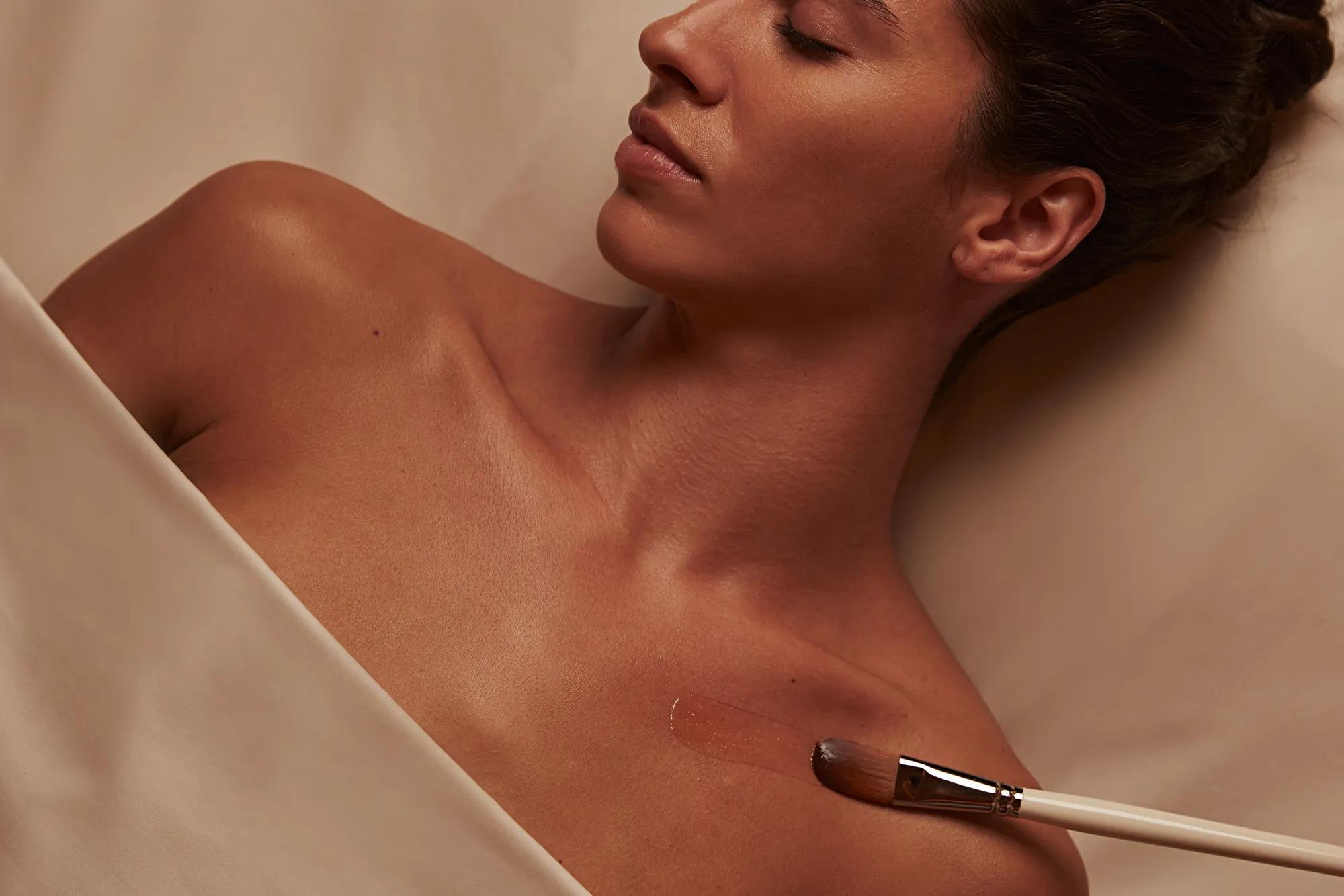 Person receiving a spa treatment, lying on their back with eyes closed, as a brush applies a soothing product to their shoulder, with soft lighting and a calm atmosphere.