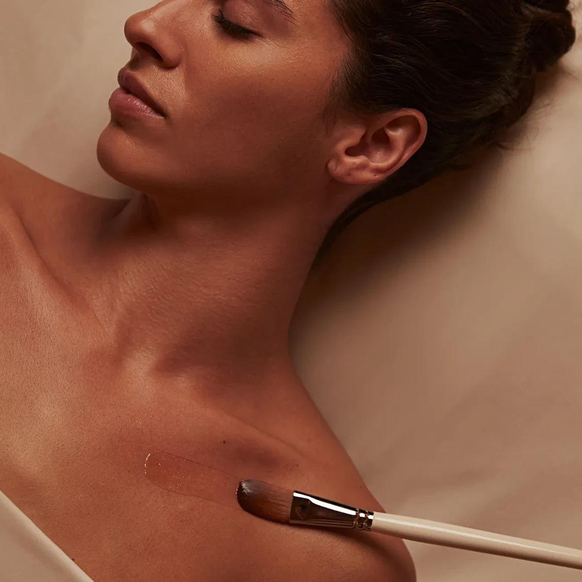 Person receiving a spa treatment, lying on their back with eyes closed, as a brush applies a soothing product to their shoulder, with soft lighting and a calm atmosphere.