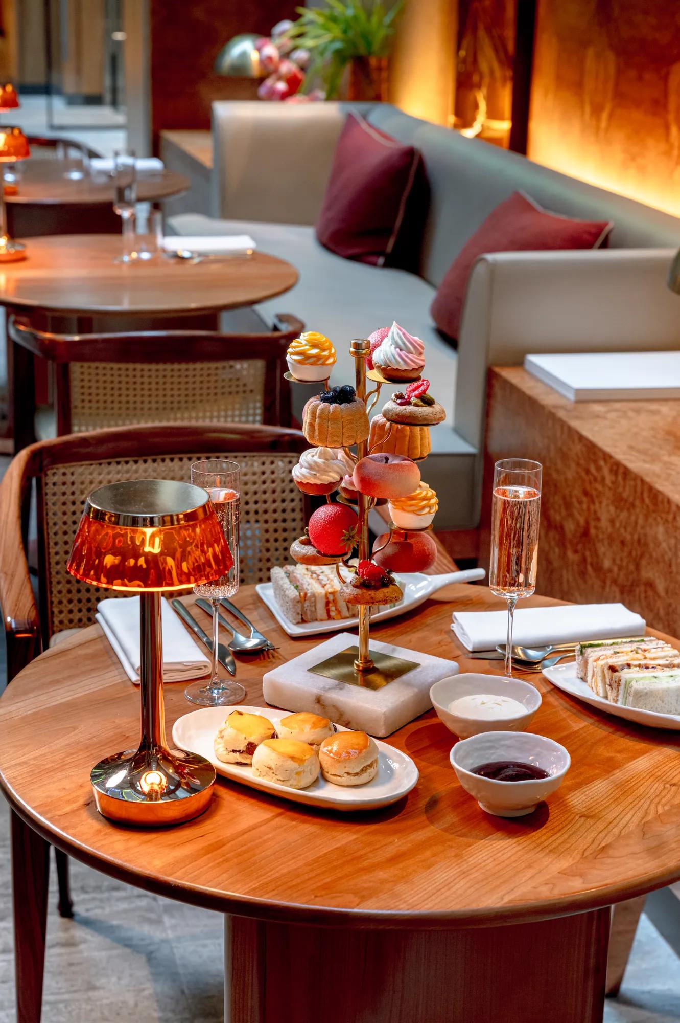 Afternoon tea spread on a wooden table with pastries, finger sandwiches, scones and champagne, set beside a lounge banquette in a warmly lit restaurant.