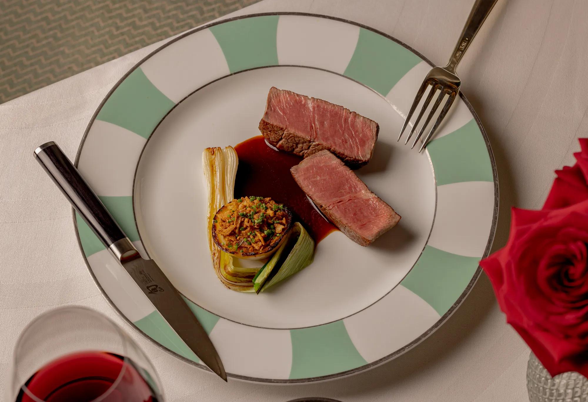 Sliced beef fillet with braised leek, stuffed onion, and red wine jus on a patterned plate.