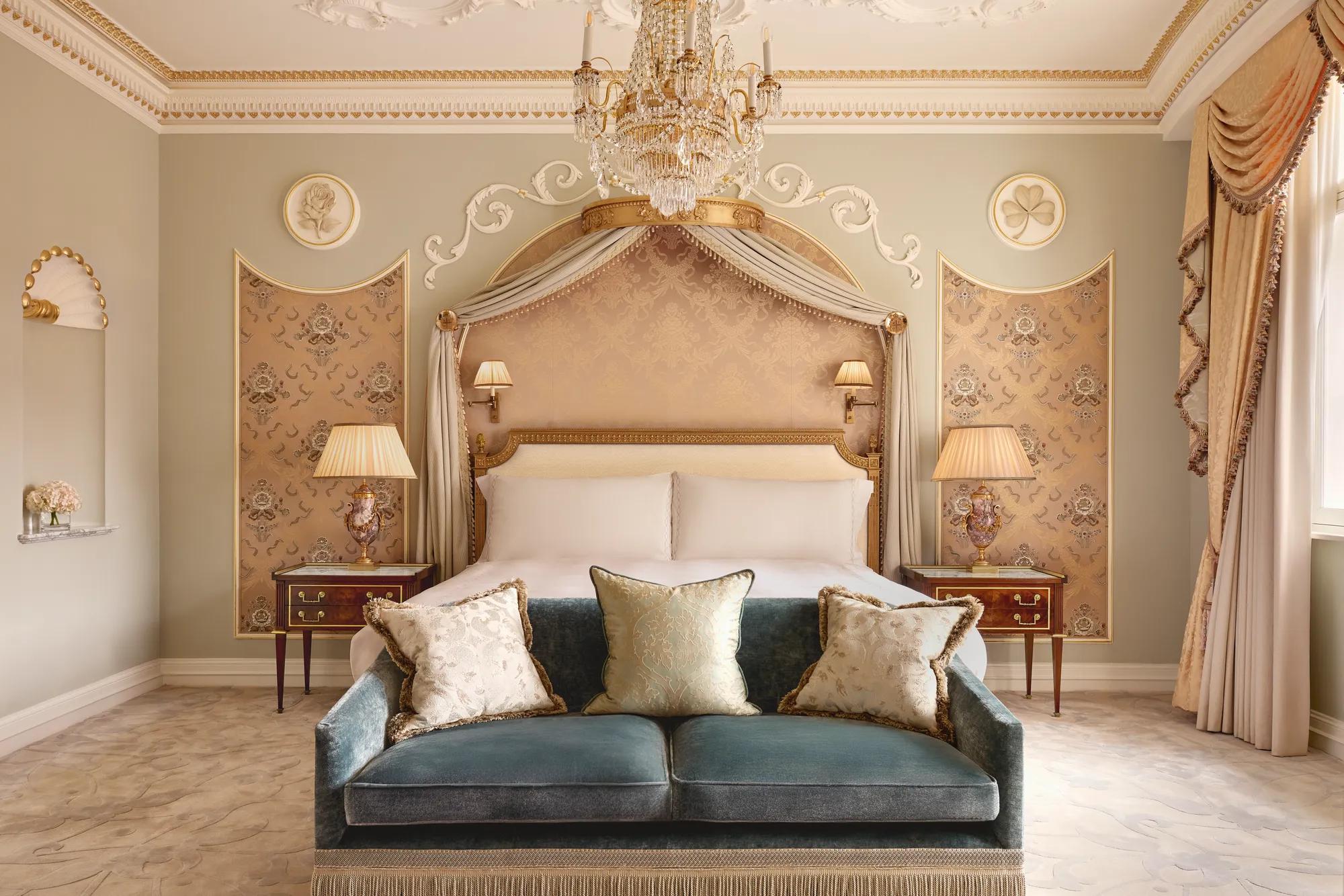 Elegant bedroom at Claridge’s with a king bed framed by a gold-accented canopy, crystal chandelier, and velvet sofa at the foot of the bed