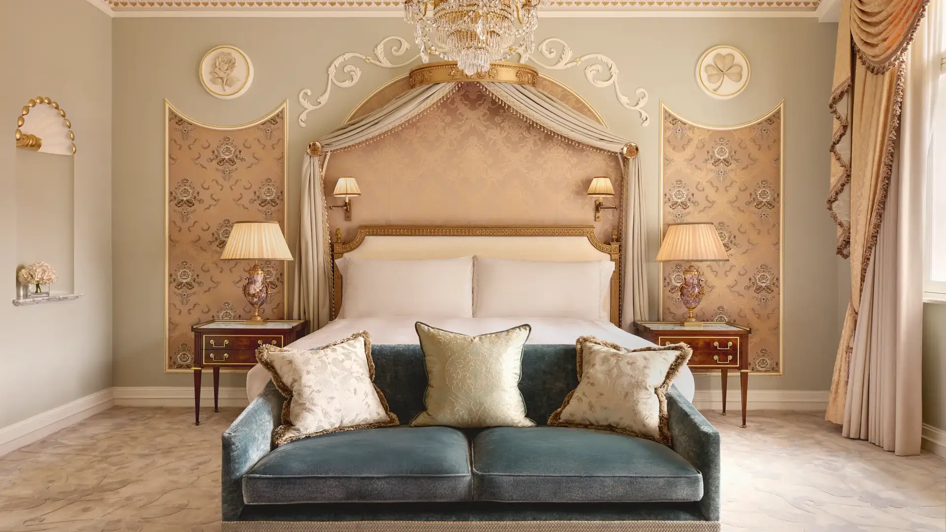 Elegant bedroom at Claridge’s with a king bed framed by a gold-accented canopy, crystal chandelier, and velvet sofa at the foot of the bed