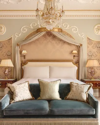 Elegant bedroom at Claridge’s with a king bed framed by a gold-accented canopy, crystal chandelier, and velvet sofa at the foot of the bed