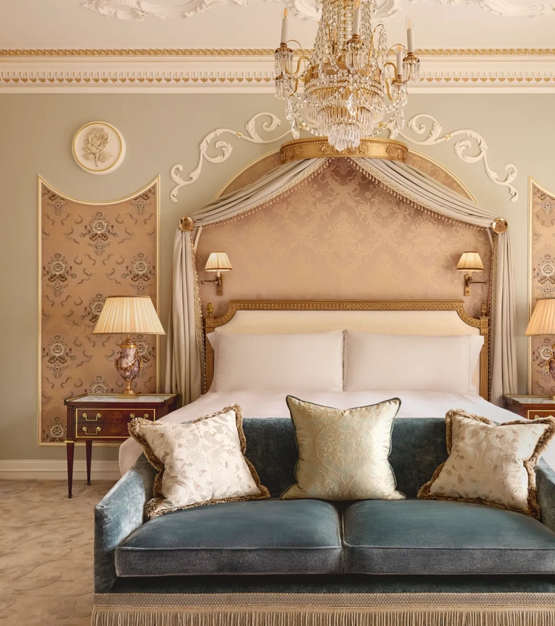 Elegant bedroom at Claridge’s with a king bed framed by a gold-accented canopy, crystal chandelier, and velvet sofa at the foot of the bed
