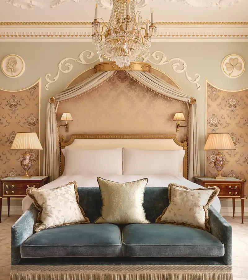 Elegant bedroom at Claridge’s with a king bed framed by a gold-accented canopy, crystal chandelier, and velvet sofa at the foot of the bed