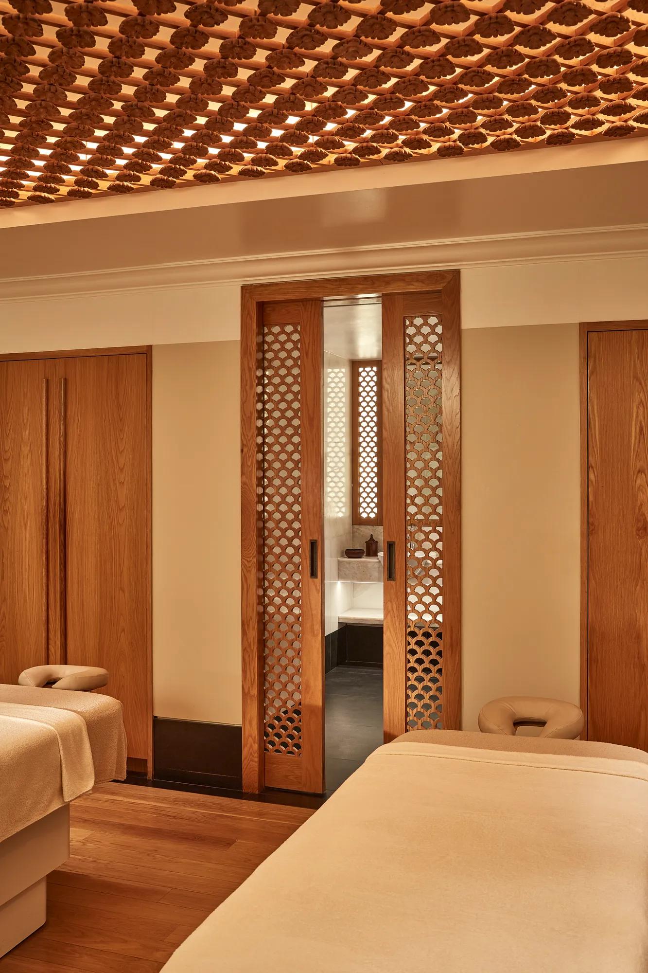 Serene spa room with two massage tables and wooden lattice doors leading to a softly lit wash area.