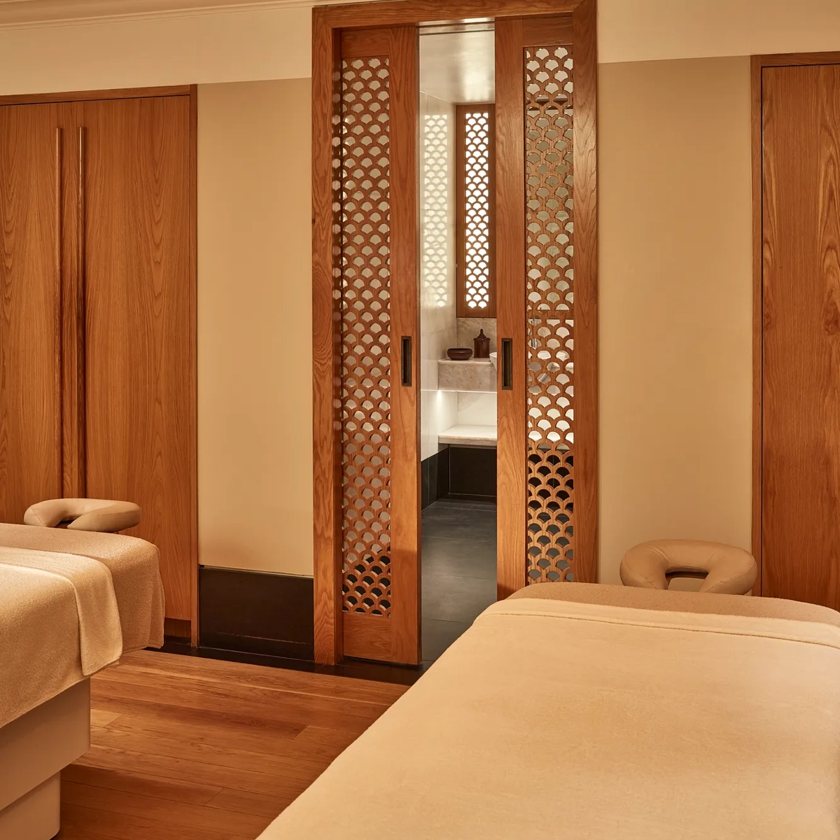 Serene spa room with two massage tables and wooden lattice doors leading to a softly lit wash area.