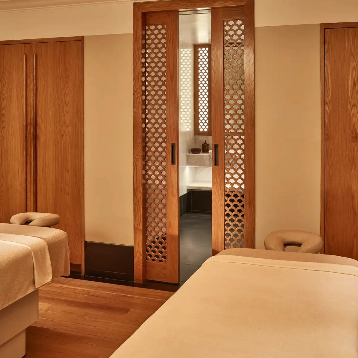 Serene spa room with two massage tables and wooden lattice doors leading to a softly lit wash area.