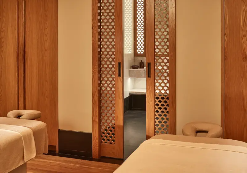 Serene spa room with two massage tables and wooden lattice doors leading to a softly lit wash area.