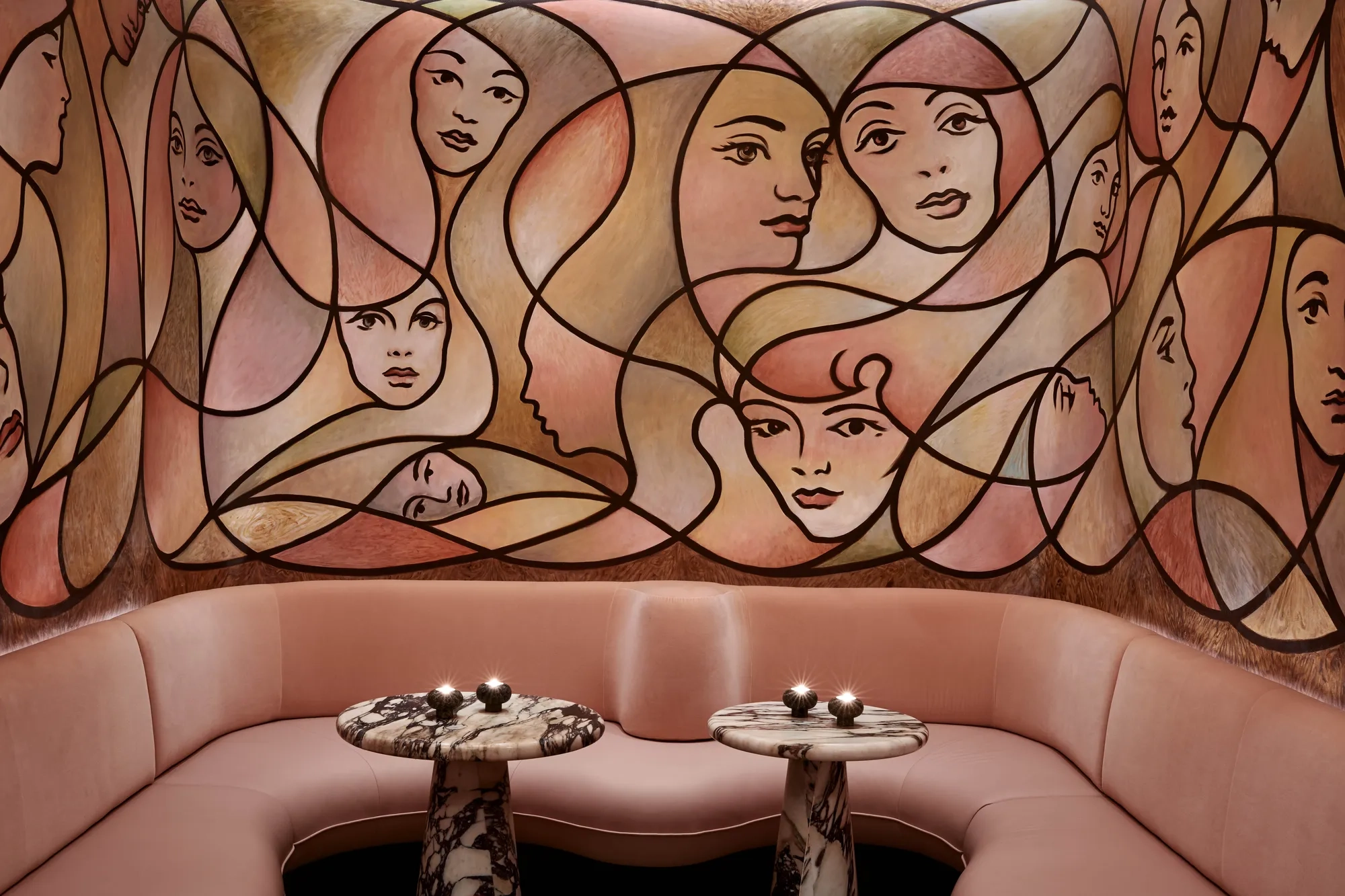 Curved blush seating with marble tables beneath large mural of abstract female faces in warm tones.