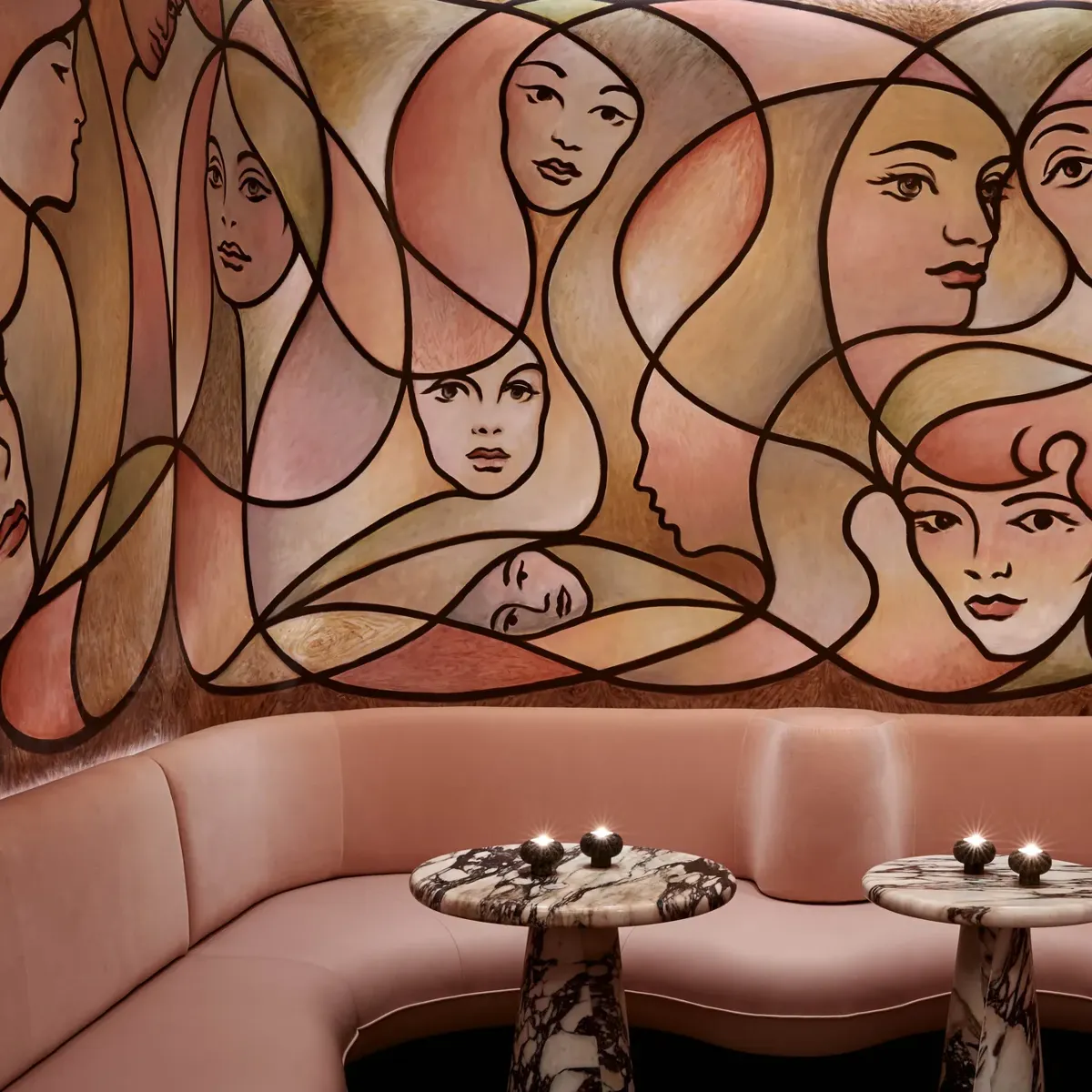 Curved blush seating with marble tables beneath large mural of abstract female faces in warm tones.