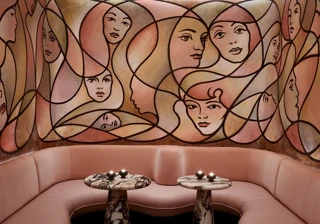 Curved blush seating with marble tables beneath large mural of abstract female faces in warm tones.