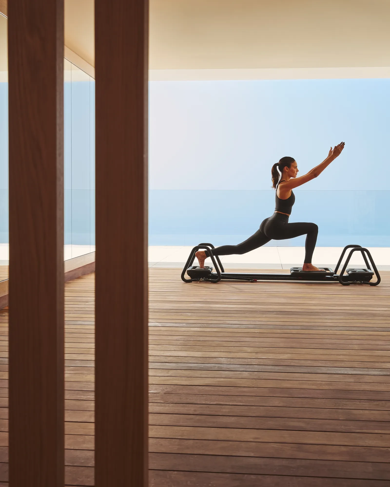 Woman performing lunging exercise on reformer machine in serene studio with panoramic sea view.