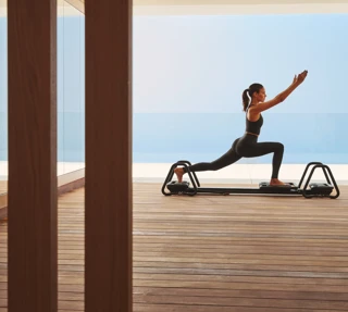 Woman performing lunging exercise on reformer machine in serene studio with panoramic sea view.