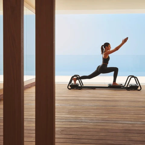 Woman performing lunging exercise on reformer machine in serene studio with panoramic sea view.