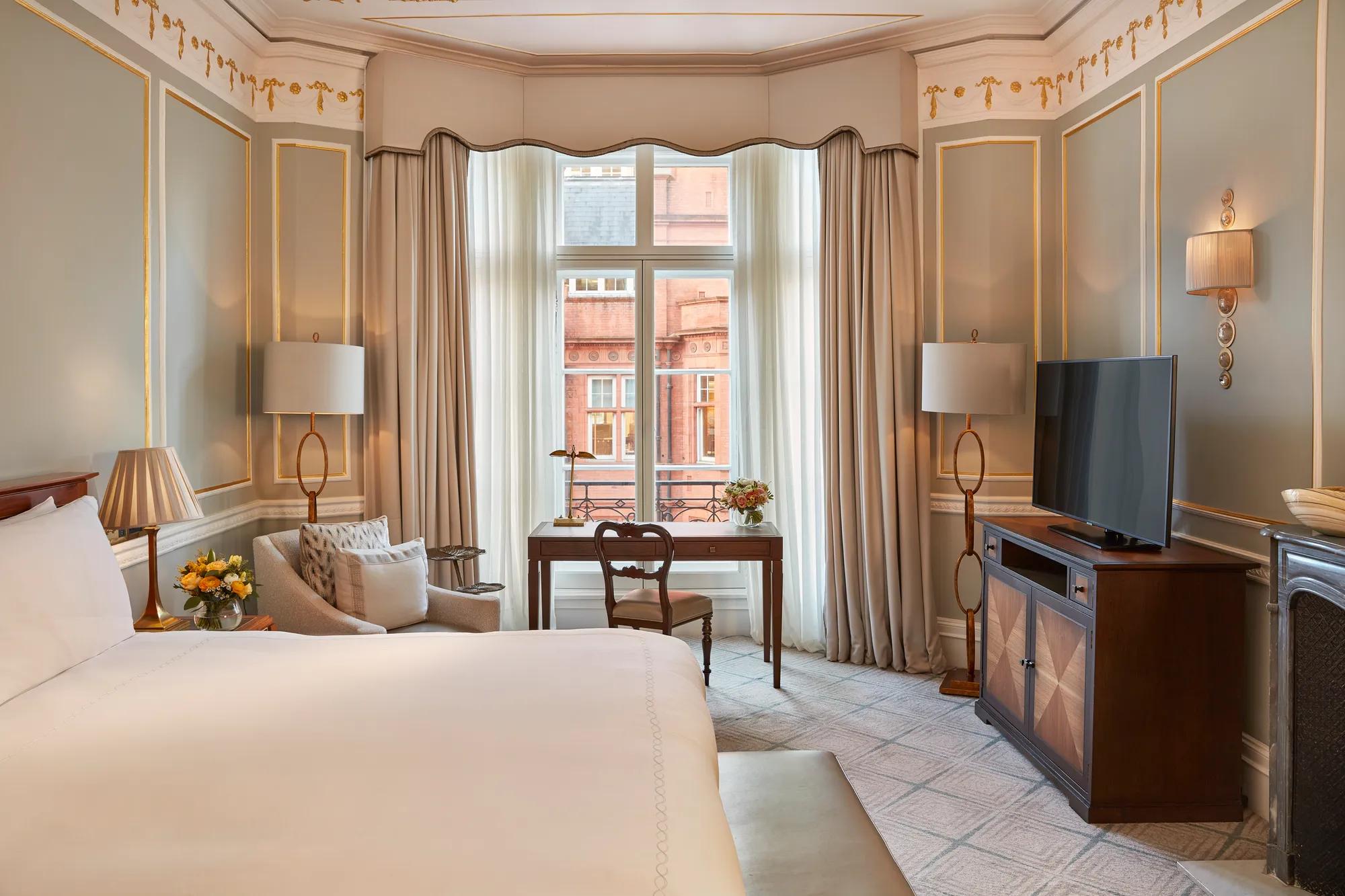 Elegant bedroom with a large bed, writing desk by a tall window, and classic décor featuring pale blue walls with gold trim and traditional furnishings.
